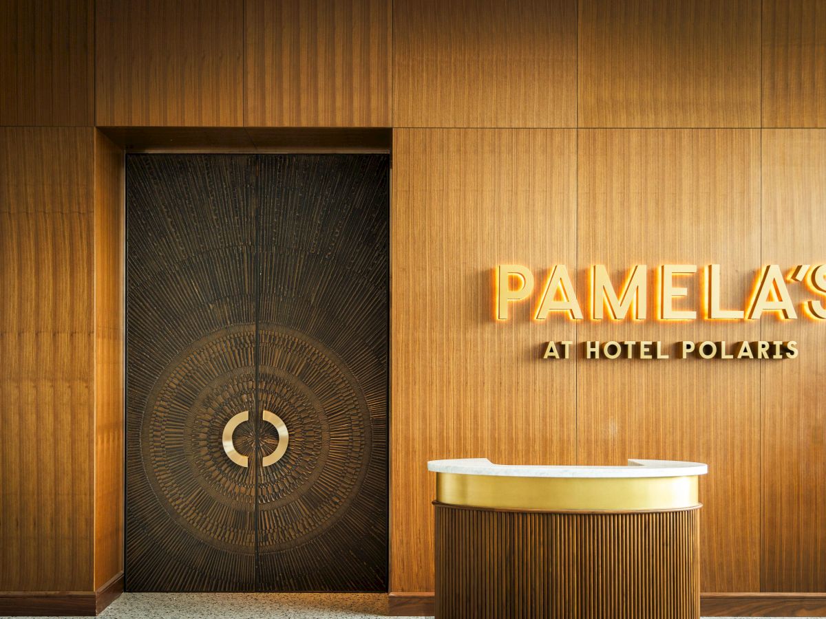 The image shows a sleek wooden interior with a door and a sign reading "PAMELA'S AT HOTEL POLARIS" above a small reception desk.