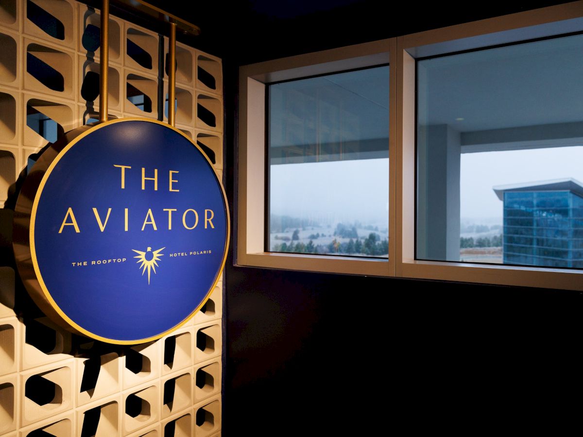 A sign for "The Aviator" lounge hangs on a patterned wall near a window with a view of a building and cloudy sky.