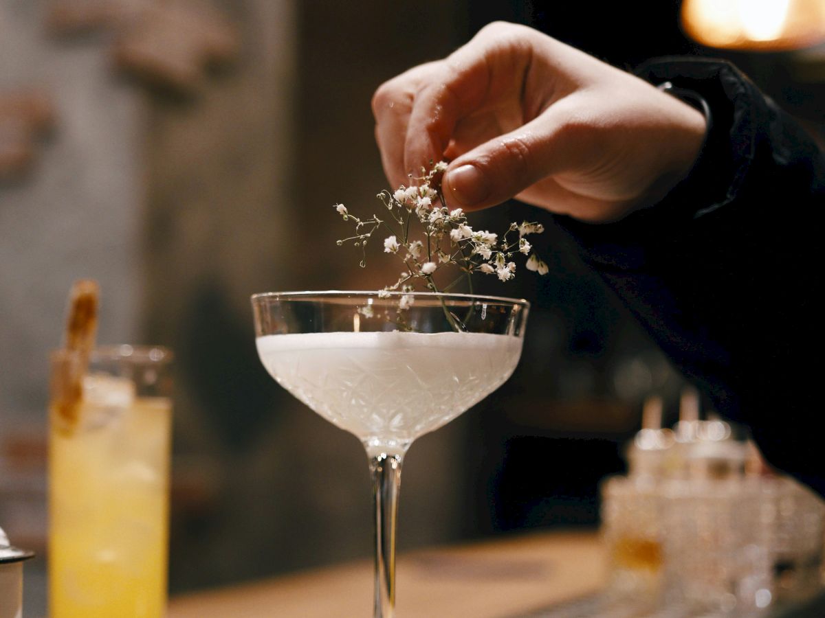 A hand is garnishing a cocktail with herbs at a bar counter. Another cocktail is placed beside it, and the bar has warm pendant lights.