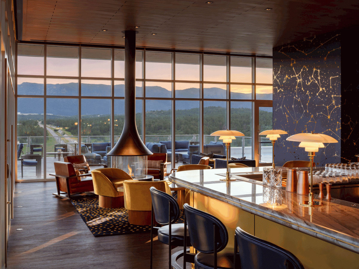 A stylish lounge with a central fireplace, bar seating, cozy chairs, large windows, and scenic mountain views at sunset.