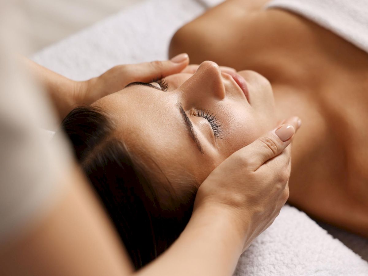 A woman receives a facial massage with eyes closed, lying on a towel, hands gently shaping her forehead.