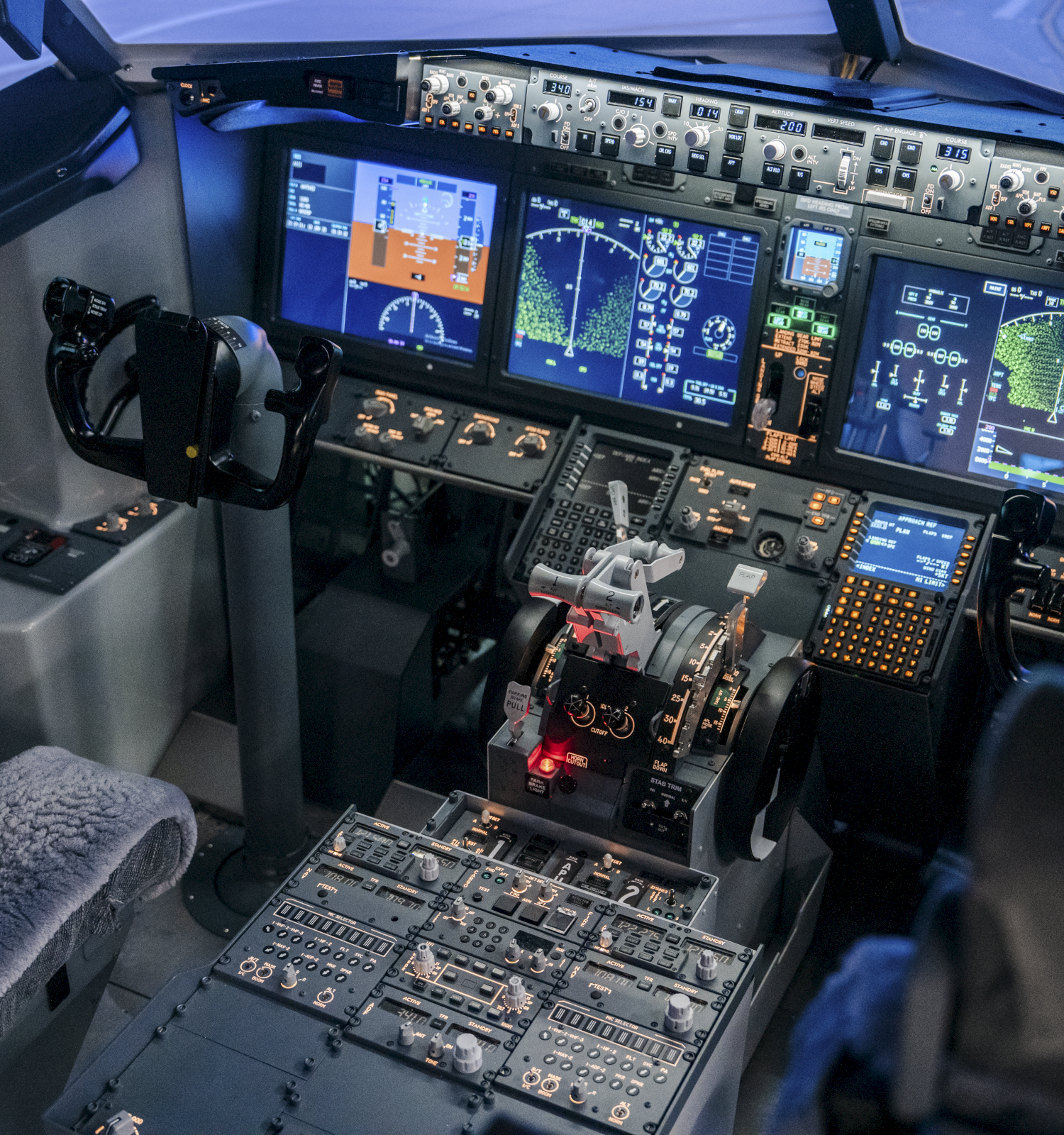 A cockpit cockpit cockpit with multiple screens, control panels, levers, and two gray seats; flight instruments and avionics dominate the panel.