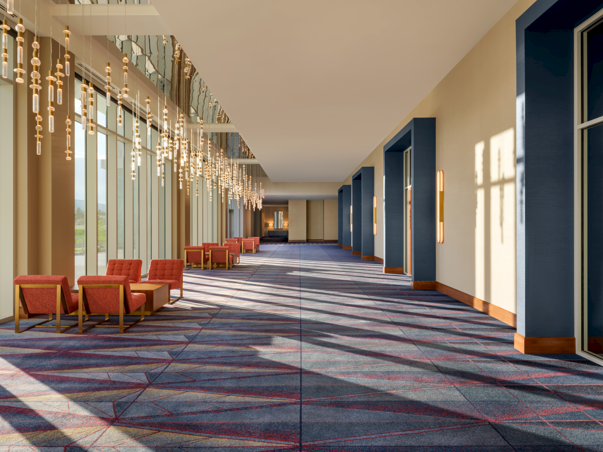 A spacious, sunlit hallway with large windows, modern chandeliers, red chairs, and patterned carpet, creating a welcoming atmosphere.