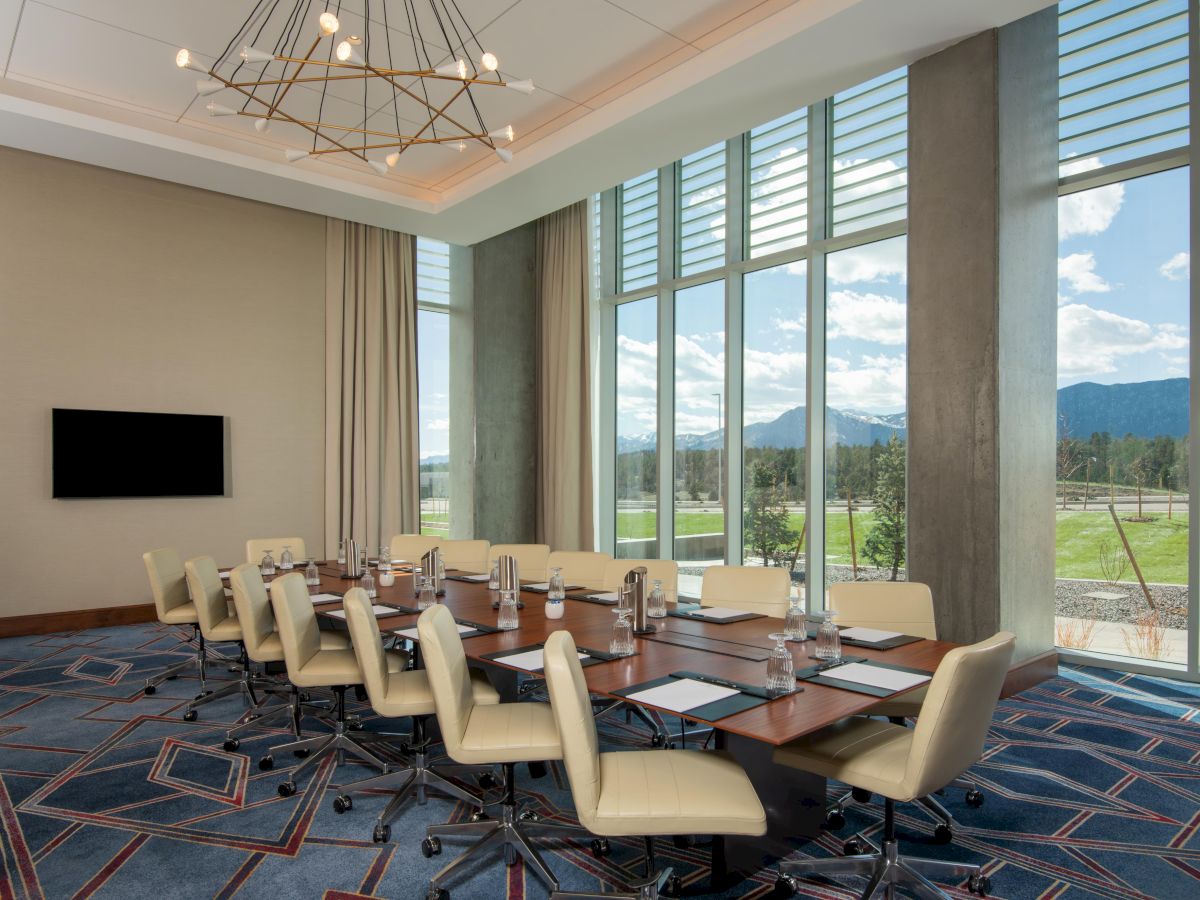 A conference room with a long table, chairs, and a wall-mounted screen, set against large windows overlooking a scenic view.