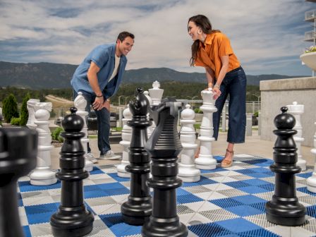 Two people play a large outdoor chess game on a blue-and-white checkered board, focused on moving the oversized pieces with a scenic backdrop.