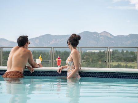 Two people in a pool, leaning on the edge with colorful drinks, chatting as mountains rise in the background&mdash;relaxed vacation vibes.
