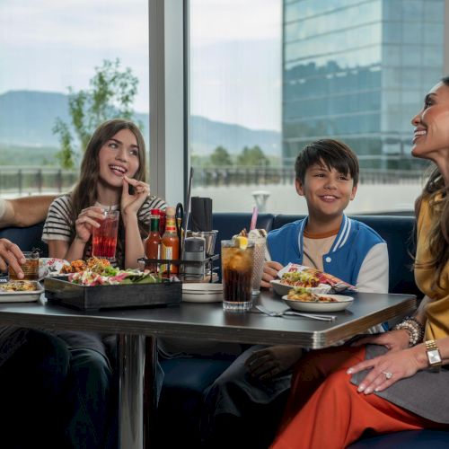 A family of four enjoys a meal at a restaurant, smiling and chatting around a table with plates, drinks, and a sunny window backdrop.
