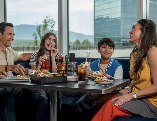 A family of four enjoys a meal at a restaurant, smiling and chatting around a table with plates, drinks, and a sunny window backdrop.