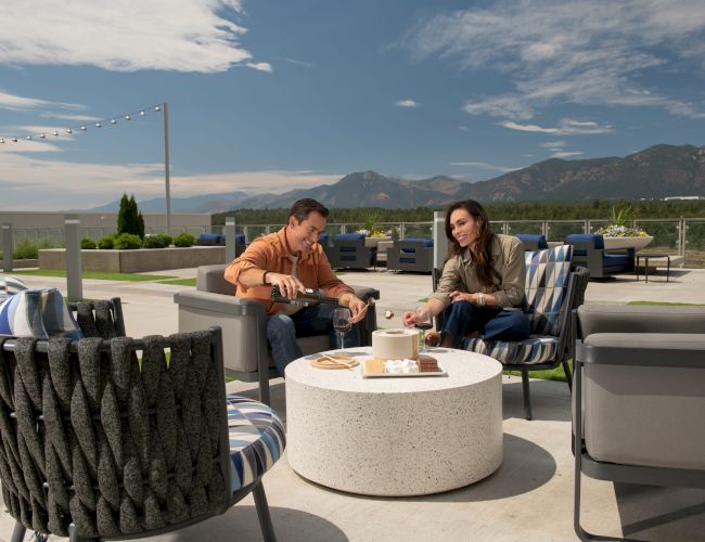 Two people sit at an outdoor lounge area with modern chairs and a round table, enjoying snacks under a clear sky with mountains in the background.