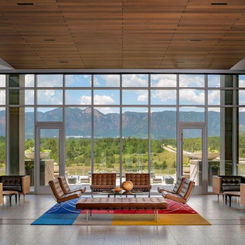A bright, modern lounge with floor-to-ceiling windows opens to a mountain view; mid-century chairs frame a colorful rug and coffee table, inviting relaxation.