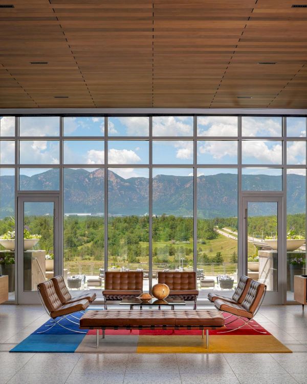 A bright, modern lounge with floor-to-ceiling windows opens to a mountain view; mid-century chairs frame a colorful rug and coffee table, inviting relaxation.