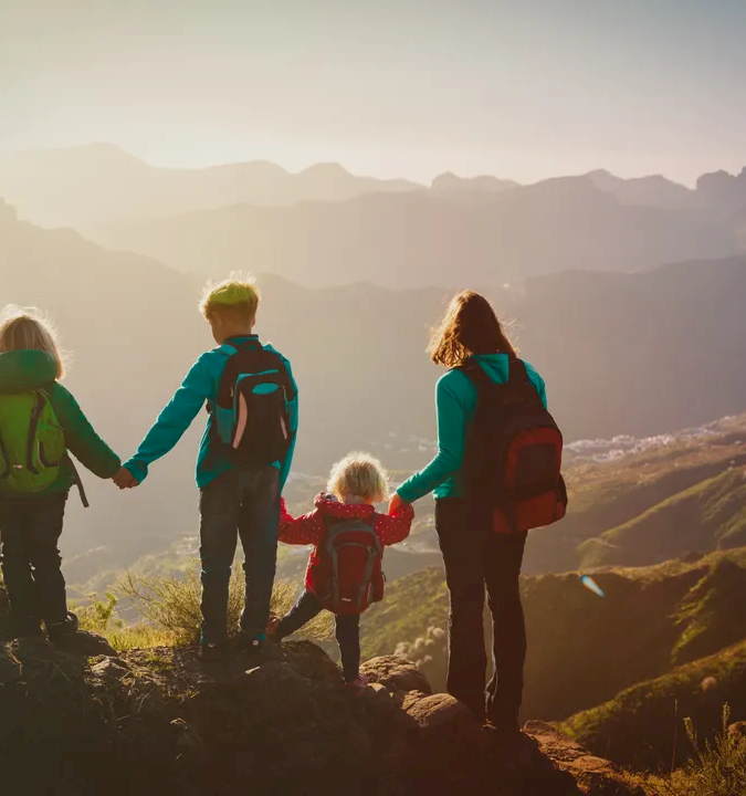 Four people (two adults, two children) hold hands at a cliff edge, backpacks on, watching a sunrise over rugged mountains.