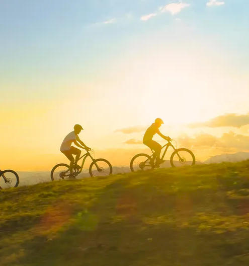 Three people riding bikes along a grassy hill at sunset, silhouettes against a warm, glowing sky as the sun sinks low.