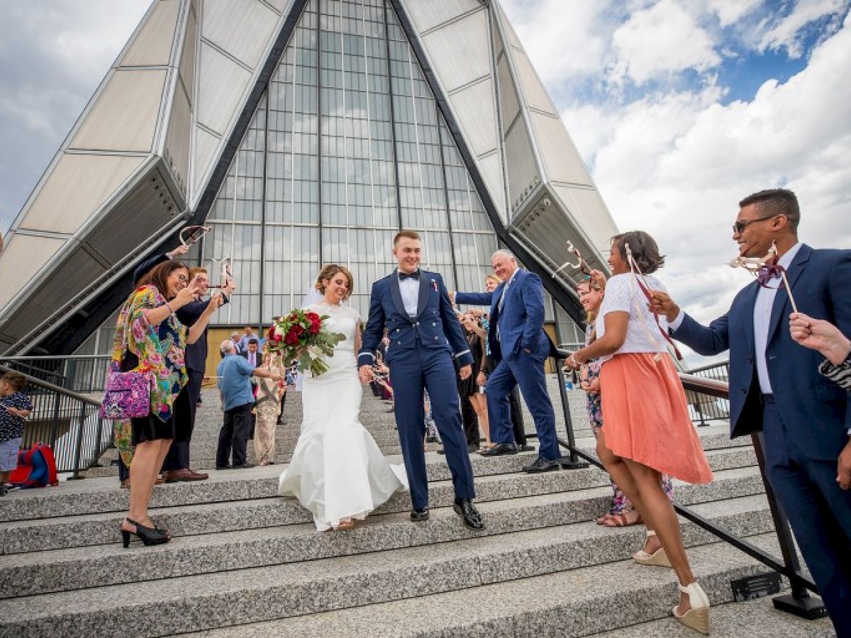 A newlywed couple exits a building, greeted by cheering guests. The bride holds a bouquet, and everyone appears joyful on the steps in front.