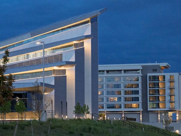A modern, angular white building with sharp lines and large glass windows sits on a grassy hill at dusk, softly illuminated.