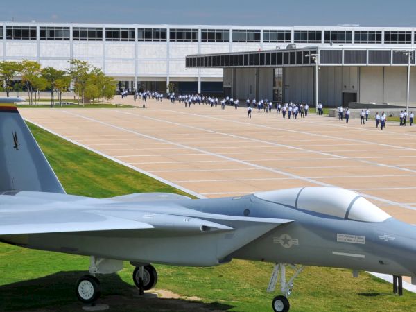 The image shows a military aircraft displayed on a grassy area in front of a large building, with people walking in the background.