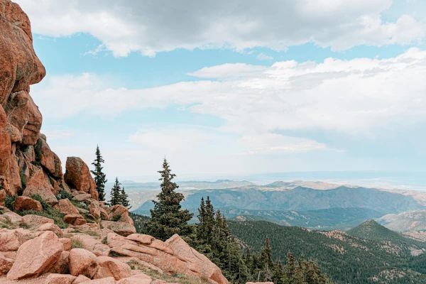 A mountain landscape with rocky cliffs, evergreen trees, and a vast view of distant hills under a partly cloudy sky, ending the sentence.