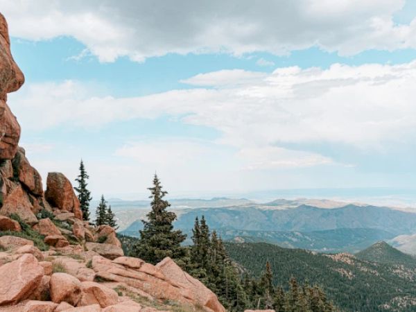 A mountain landscape with rocky cliffs, evergreen trees, and a vast view of distant hills under a partly cloudy sky, ending the sentence.