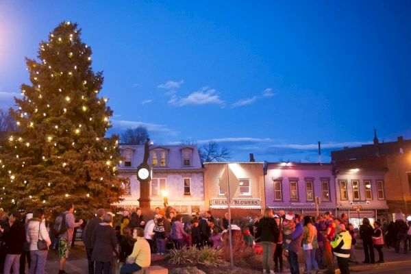 A crowd gathers around a large, decorated Christmas tree in a town square, with buildings and a clock in the background at dusk.