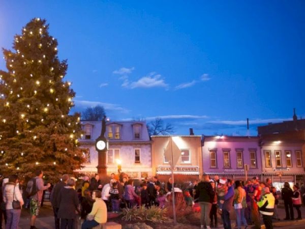 A crowd gathers around a large, decorated Christmas tree in a town square, with buildings and a clock in the background at dusk.