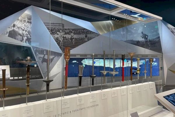 The image shows a museum exhibit with various Olympic torches displayed in a glass case. Historical photos are visible in the background.