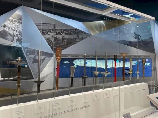 The image shows a museum exhibit with various Olympic torches displayed in a glass case. Historical photos are visible in the background.