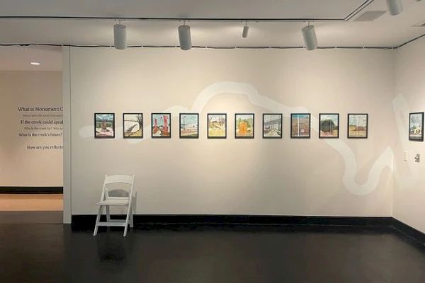 A row of framed pictures is displayed on a white wall in a gallery. There's also a white chair in front of the wall.