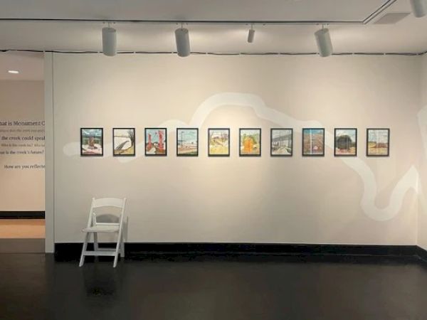 A row of framed pictures is displayed on a white wall in a gallery. There's also a white chair in front of the wall.