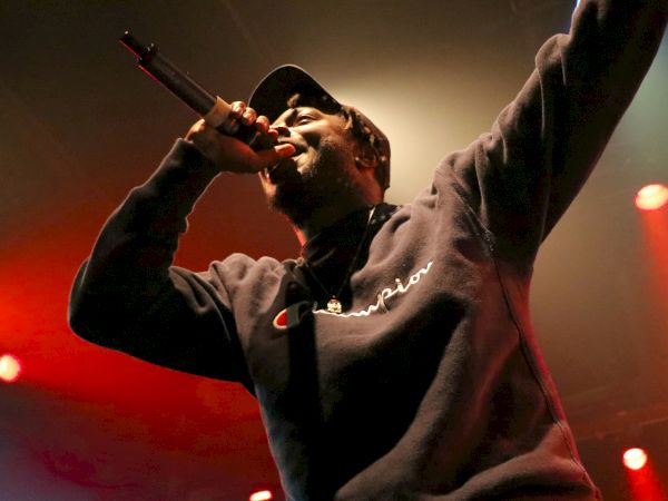 A person is performing on stage, holding a microphone, wearing a Champion sweatshirt, with red and blue lighting in the background.