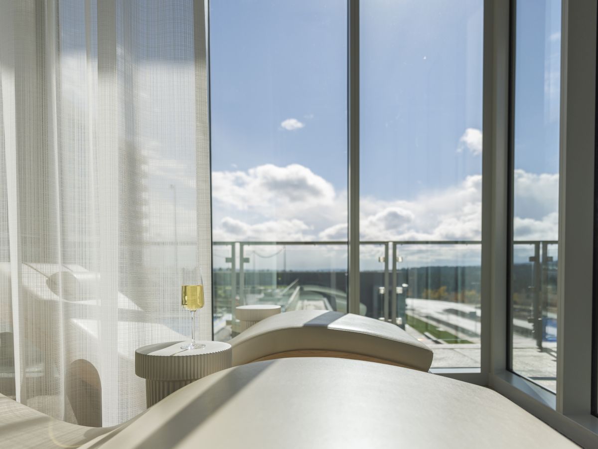 A modern hotel room scene with a bed, a champagne glass on a side table, and curtains framing a blue sky view.