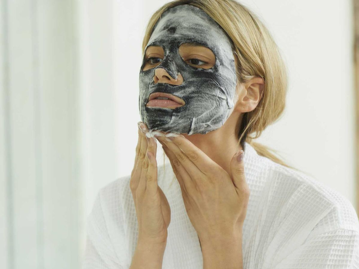 A woman with a gray facial mask is applying it to her face, wearing a white robe, in a bright bathroom.