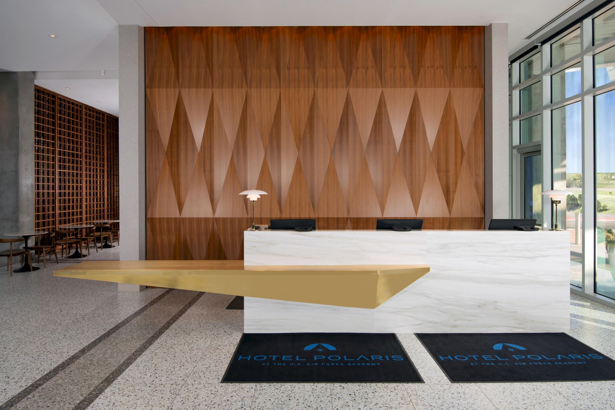 A hotel reception area with a modern design, featuring a wood panel wall, two desks, lamps, and "Hotel Polaris" mats on the floor.