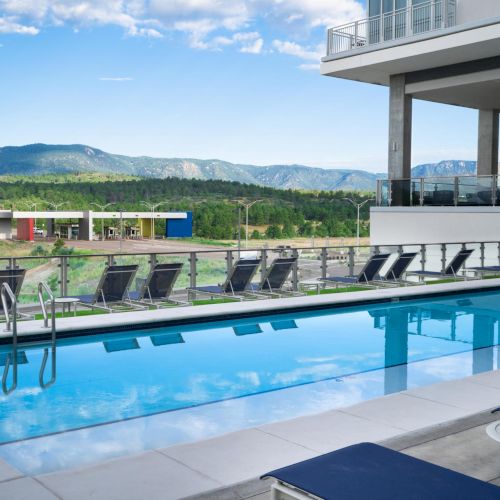 A modern poolside area with a long rectangular swimming pool, lounge chairs along the deck, and a glass-walled building upstairs; hills in the distance. Keep it concise.