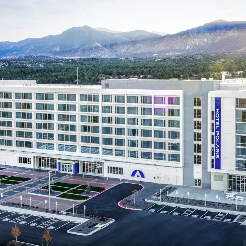 A large multi-story hospital building with a mountain backdrop, surrounded by parking areas and greenery under a clear sky.