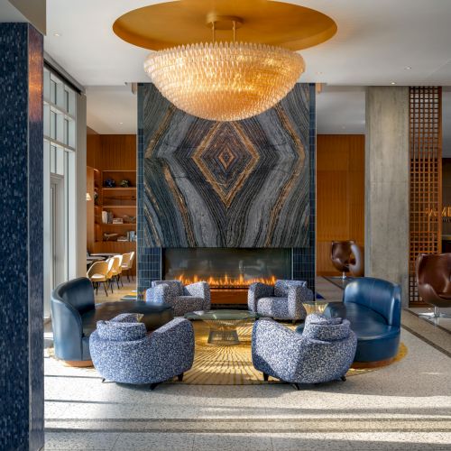 A modern lobby with two blue armchairs facing a round coffee table, a large circular light fixture above, and a decorative wall panel behind.