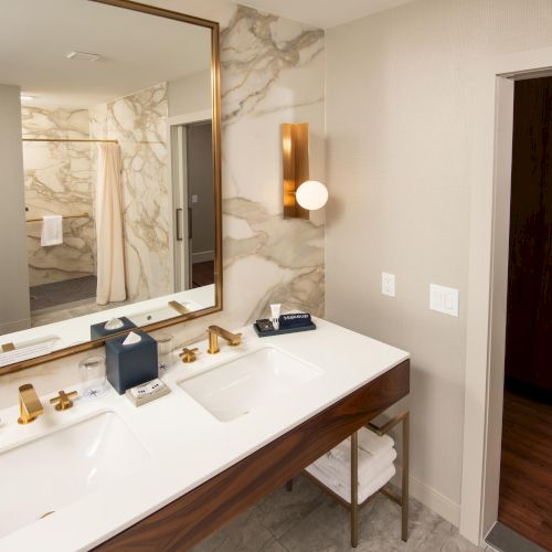 The image shows a modern bathroom with a double sink vanity, marble walls, gold fixtures, and bathrobes hanging in an open closet.
