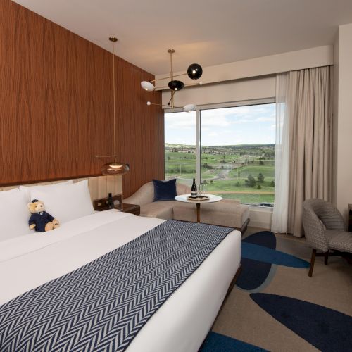 A modern hotel room with a plush bed, a teddy bear, a desk, a cozy sitting area by the window, and a scenic view outside.