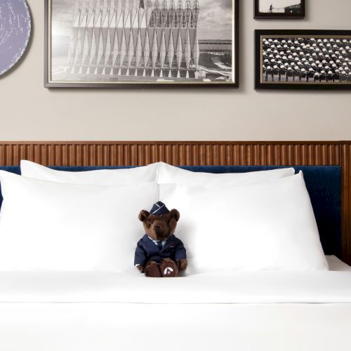 A teddy bear in a uniform sits on a bed with white pillows; framed pictures are on the wall above it.