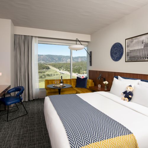 A modern hotel room with a large bed, sofa, desk, and window view. Decor includes photos, a teddy bear, and wine on the table.