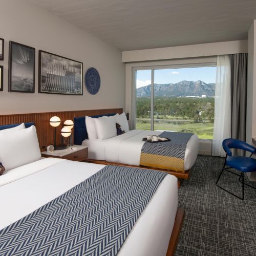 A modern hotel room with two beds, wall art, a window with a mountain view, and a desk with a chair.