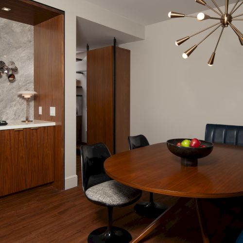 A modern dining area with a wooden table, black chairs, wall art, a hanging light fixture, and a decor bowl with apples.