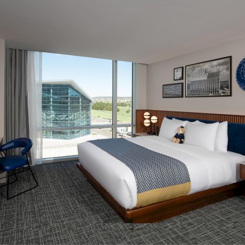 A modern hotel room features a large bed, stylish lighting, a desk, and a view of a glass building through a window.