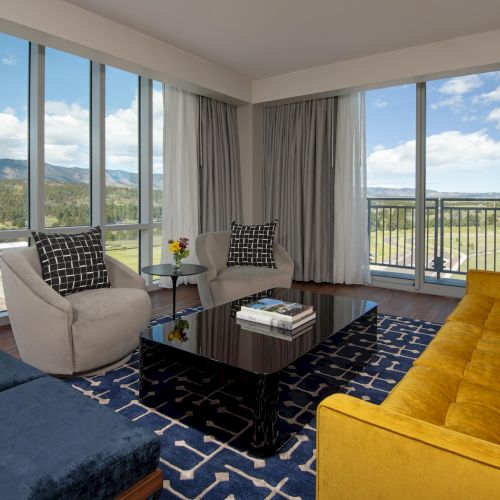 A modern living room with large windows, offering a scenic view, features a mustard sofa, two chairs, a dark coffee table, and a patterned rug.