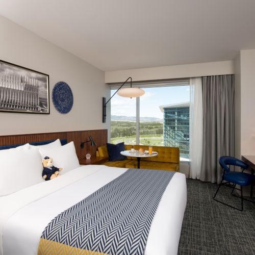 A modern hotel room with a bed, yellow sofa, desk, and window view. Art hangs on the wall, and a teddy bear sits on the bed.