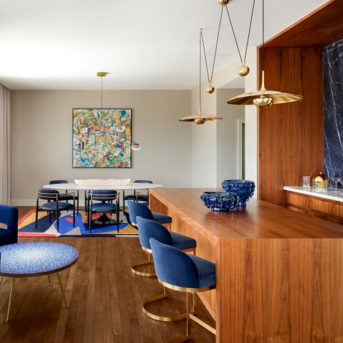 A modern kitchen and dining area with a wooden island, blue chairs, pendant lights, a marble backsplash, and a colorful wall art piece on a beige wall.