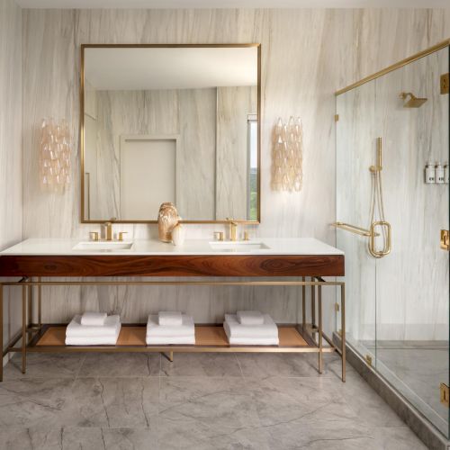 A luxurious bathroom with a double vanity, gold fixtures, a large mirror, marble floors, and a glass-walled shower, all in soft neutral tones.