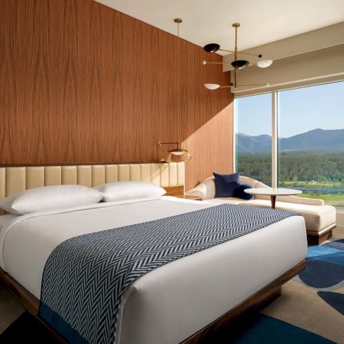 A modern hotel room with a king bed, wooden accent wall, plush seating, and a large window overlooking mountains and a valley, bright and airy.