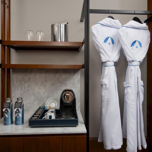 The image shows a hotel setup with two bathrobes, a coffee machine, coffee pods, bottled drinks, glasses, and an ice bucket on shelves.