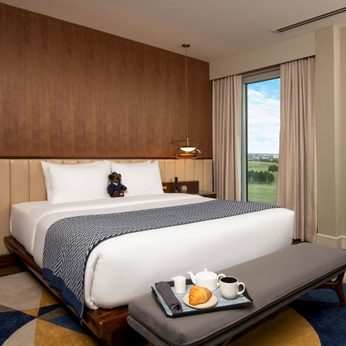 A hotel room with a modern design, featuring a king-size bed, a teddy bear, breakfast on a bench, and a view of a green landscape.
