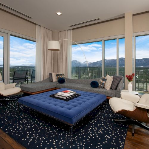 A modern living room with a large white chair, blue ottoman, sleek sofa, and a mountain view through the windows, decorated with books and a vase.